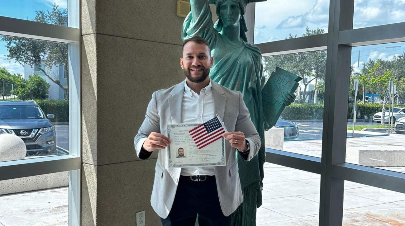 2 Bearded man in a gray blazer holding a U.S. citizenship certificate and small American flag beside a Statue of Liberty statue