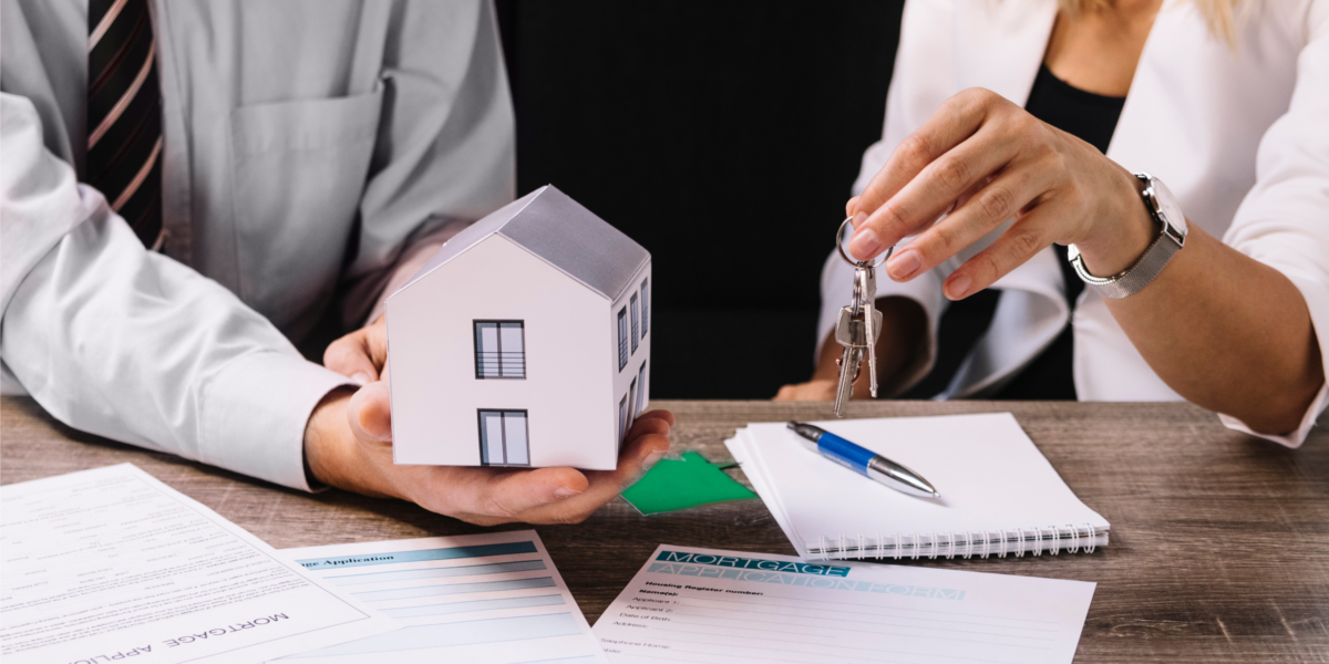 1 A person handing over house keys to another holding a model house over paperwork